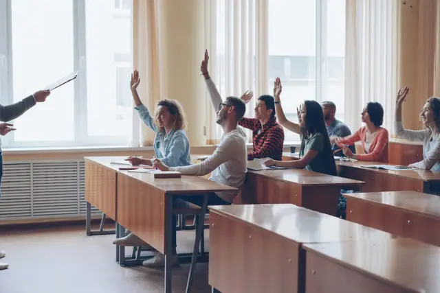 Students seated around a seminar table raising their hands towards a teacher.