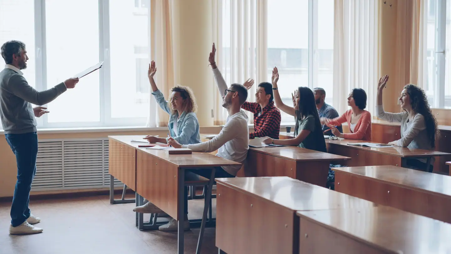 Students seated around a seminar table raising their hands towards a teacher.