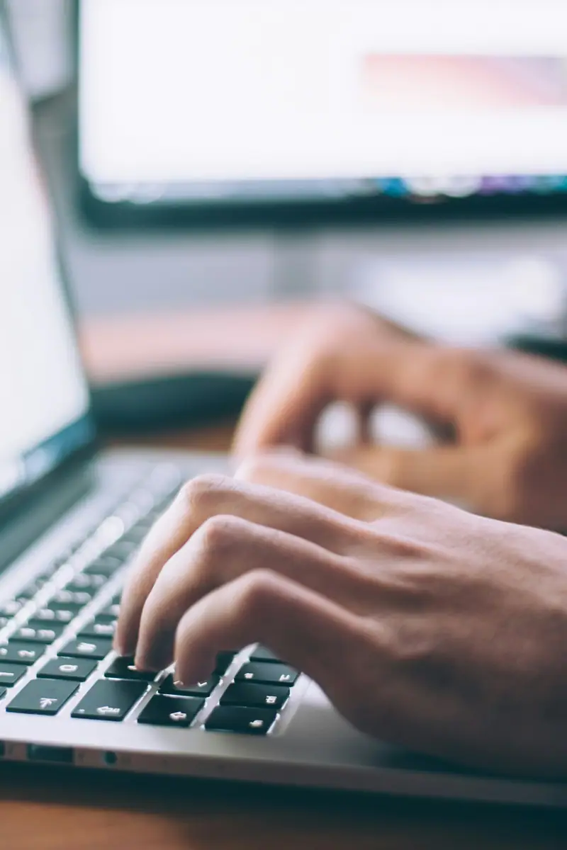 Hands typing on a laptop keyboard beside a second screen.
