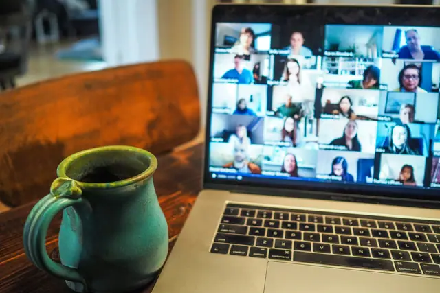 A laptop on a wooden table showing a grid of video-call participants, a mug beside it.